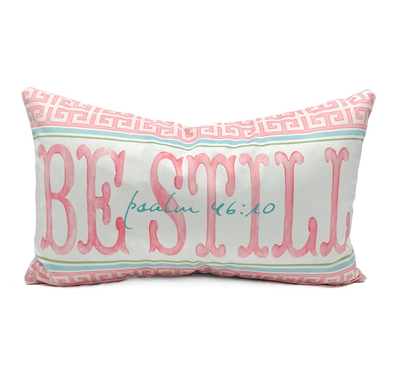 May include: A white rectangular pillow with a pink and green Greek key pattern border. The pillow has the words "BE STILL" in pink lettering with the words "psalm 46:10" in teal lettering below.