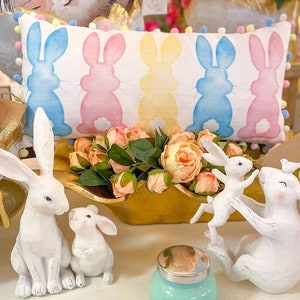 May include: A decorative Easter-themed display featuring a rectangular pillow with bunny silhouettes in blue, pink, and yellow. White bunny figurines and a teal jar with a silver lid are also present, along with a gold-colored wooden bowl filled with pink flowers.