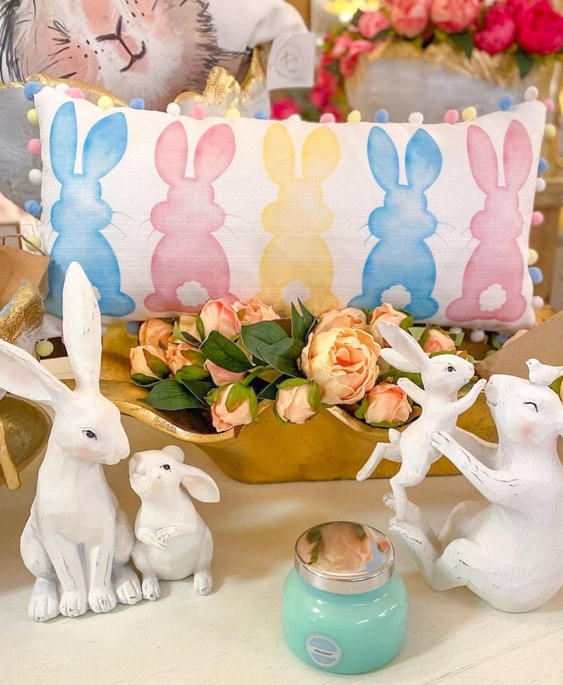 May include: A decorative Easter-themed display featuring a rectangular pillow with bunny silhouettes in blue, pink, and yellow. White bunny figurines and a teal jar with a silver lid are also present, along with a gold-colored wooden bowl filled with pink flowers.
