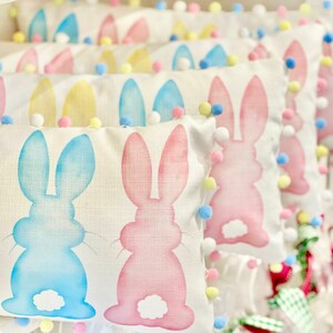 May include: Rectangular Easter-themed pillows with bunny designs in pastel blue and pink. Each pillow is trimmed with colorful pom-poms. The pillows are arranged in a row, creating a festive and decorative display.