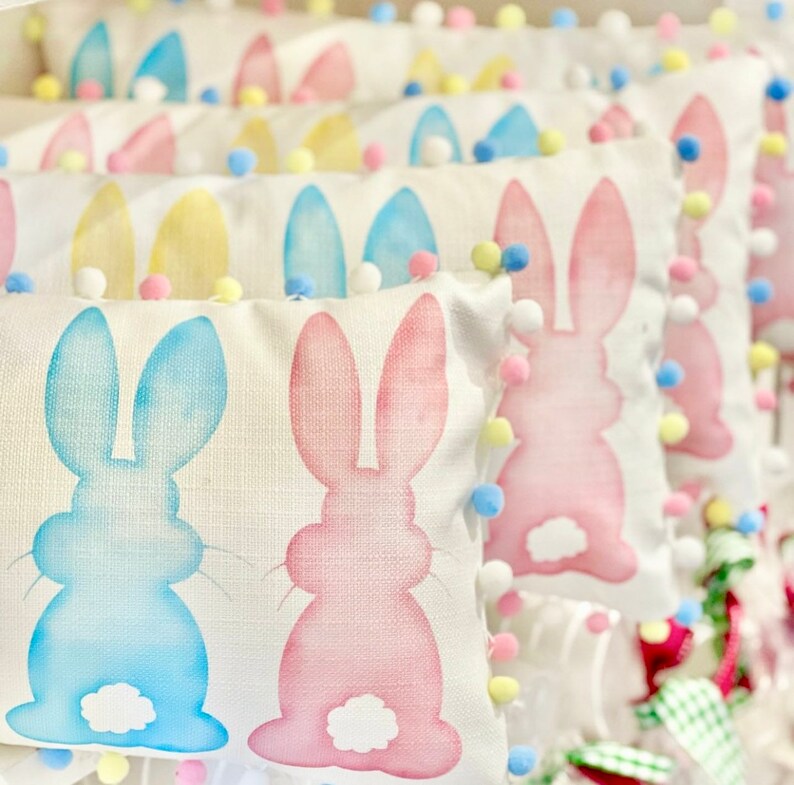 May include: Rectangular Easter-themed pillows with bunny designs in pastel blue and pink. Each pillow is trimmed with colorful pom-poms. The pillows are arranged in a row, creating a festive and decorative display.