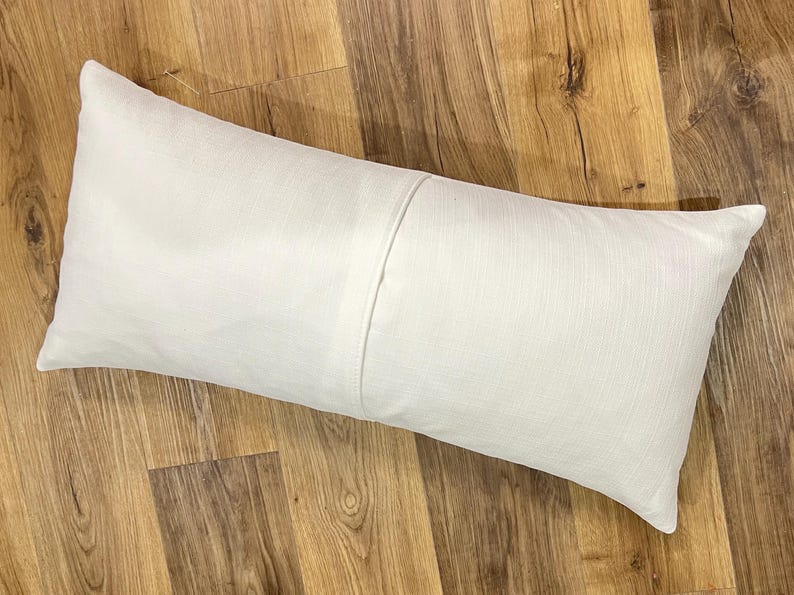 K&ouml;nnte beinhalten: Ein rechteckiger Lendenkissenbezug in einem einfarbigen Off-White. Der Stoff weist eine sichtbare Webstruktur auf und hat eine vertikale Naht, die mittig &uuml;ber das Kissen verl&auml;uft. Das Design ist schlicht und funktional mit einer neutralen Optik.