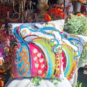 May include: A square throw pillow featuring a vibrant, multi-colored painted pumpkin design on a white background. The pumpkin is decorated with bright blue, hot pink, lime green, and gold metallic accents, including swirling vines and dots.