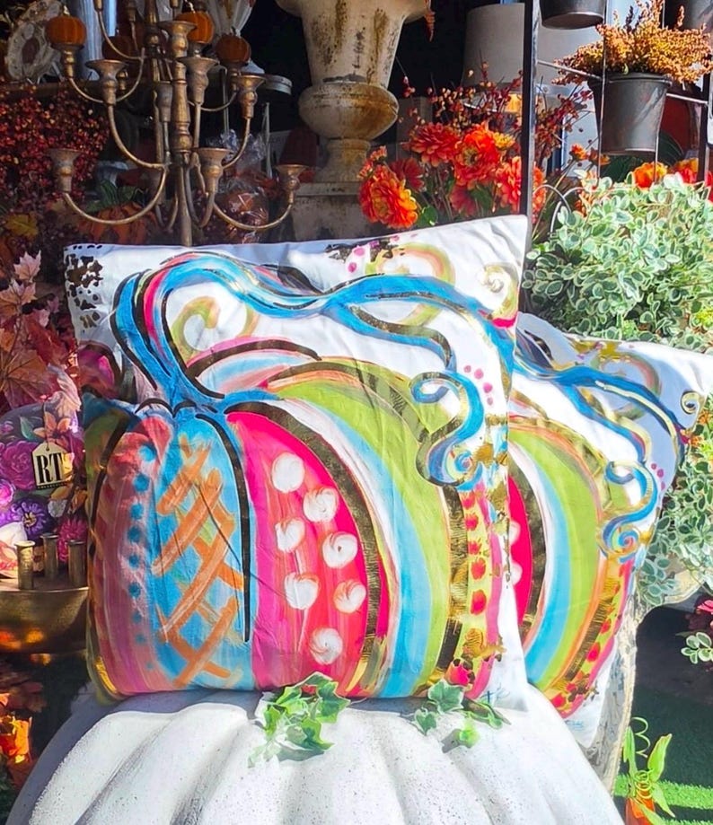 May include: A square throw pillow featuring a vibrant, multi-colored painted pumpkin design on a white background. The pumpkin is decorated with bright blue, hot pink, lime green, and gold metallic accents, including swirling vines and dots.