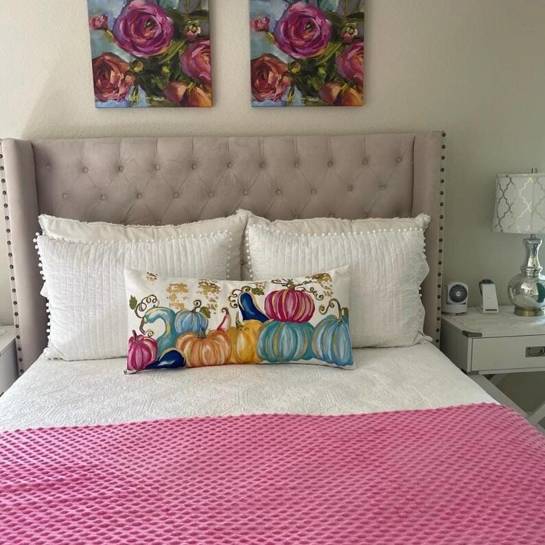 May include: A white bed with a pink quilted bedspread. The bed has a white headboard with tufted buttons. There are two white pillows with white lace trim and a decorative pillow with a colourful pumpkin design.