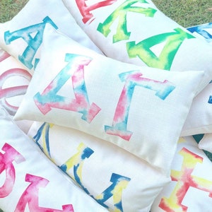 May include: A collection of white pillow covers with tie-dye letters. The letters are in various colors, including pink, blue, yellow, and green. The pillows are arranged in a pile, with the letters facing up.