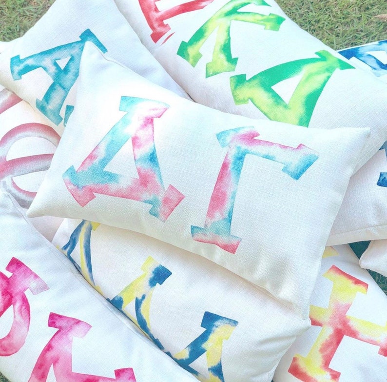 May include: A collection of white pillow covers with tie-dye letters. The letters are in various colors, including pink, blue, yellow, and green. The pillows are arranged in a pile, with the letters facing up.