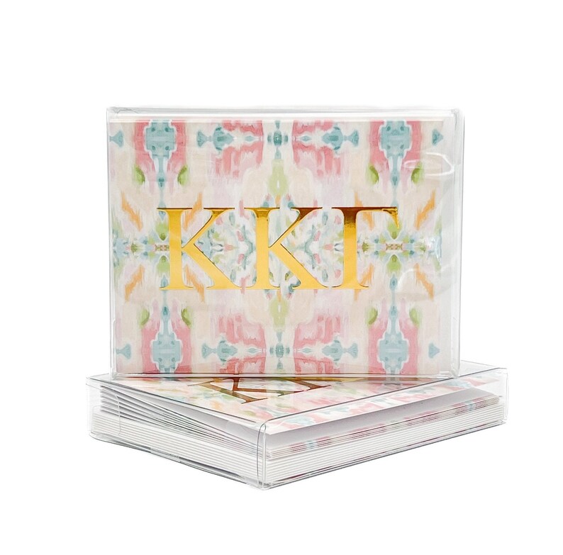 K&ouml;nnte beinhalten: Ein Set mit Notizbl&auml;ttern in einer Box mit einem pinkfarbenen, blauen und gr&uuml;nen Ikat-Muster und goldenen Folienbuchstaben, die "KK&Gamma;" lesen.