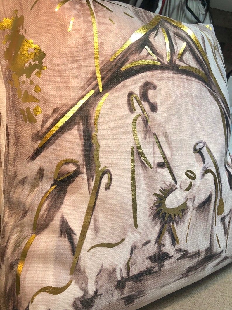 May include: A beige throw pillow with a gold and brown illustration of the nativity scene. The illustration features a star, a manger, and three figures.