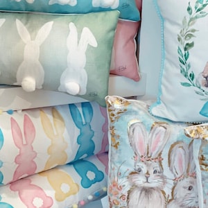 May include: A collection of decorative Easter-themed pillows. Pillows feature bunny designs, including bunny tails, silhouettes, and watercolor illustrations of bunnies with floral crowns. Colors include pastel blues, greens, pinks, and yellows.