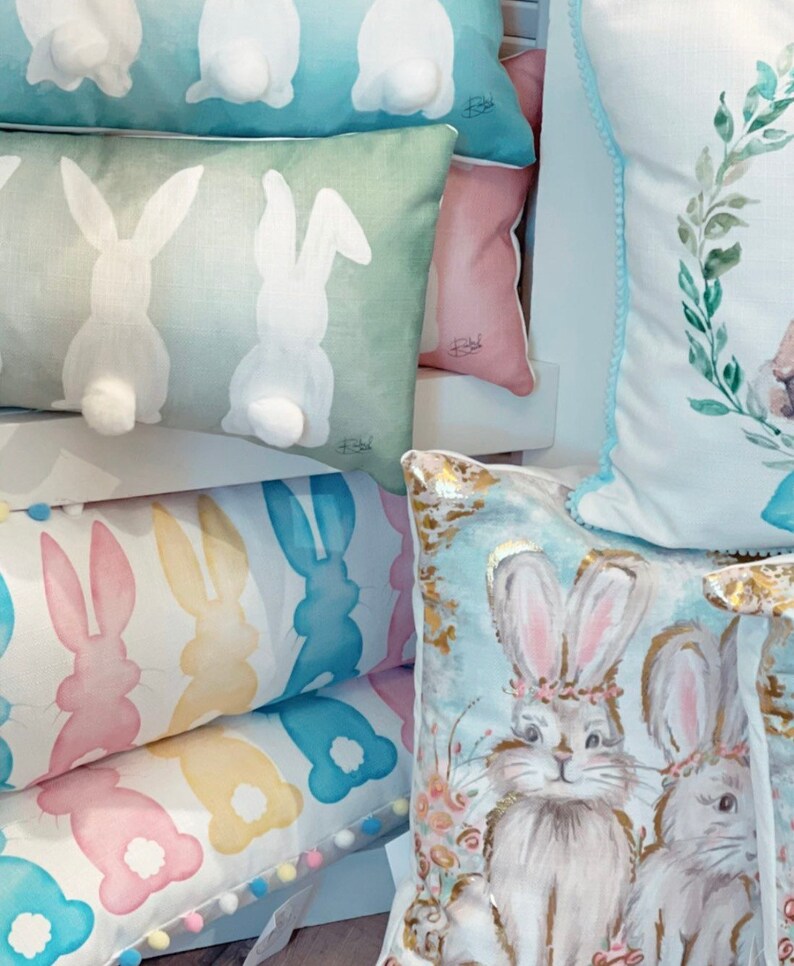 May include: A collection of decorative Easter-themed pillows. Pillows feature bunny designs, including bunny tails, silhouettes, and watercolor illustrations of bunnies with floral crowns. Colors include pastel blues, greens, pinks, and yellows.