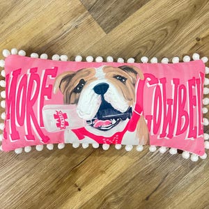 K&ouml;nnte beinhalten: Ein rechteckiges, rosafarbenes Dekokissen mit wei&szlig;er Bommelborte. In der Mitte ist eine braun-wei&szlig;e Bulldogge abgebildet, die eine Kuhglocke im Maul h&auml;lt. Der Text MORE COWBELL ist in gro&szlig;en, fetten rosa Buchstaben auf der Vorderseite gedruckt.