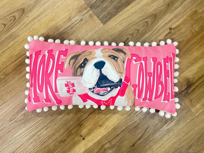 K&ouml;nnte beinhalten: Ein rechteckiges, rosafarbenes Dekokissen mit wei&szlig;er Bommelborte. In der Mitte ist eine braun-wei&szlig;e Bulldogge abgebildet, die eine Kuhglocke im Maul h&auml;lt. Der Text MORE COWBELL ist in gro&szlig;en, fetten rosa Buchstaben auf der Vorderseite gedruckt.