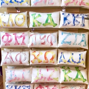 May include: A collection of 12 white pillow covers with colorful tie-dye designs featuring Greek letters. The letters include:  &Alpha;&Delta;&Pi;, &Kappa;&Delta;&Kappa;, &Kappa;&Kappa;&Gamma;, &Phi;&Mu;, &Tau;&Beta;&Phi;, &Zeta;&Tau;&Alpha;, &Alpha;&Phi;&Phi;, &Chi;&Omega;, &Delta;&Delta;&Delta;, &Delta;&Sigma;&Zeta;, &Kappa;&Delta;&Kappa;, &Kappa;&Kappa;&Gamma;, &Phi;&Mu;, &Tau;&Beta;&Phi;.