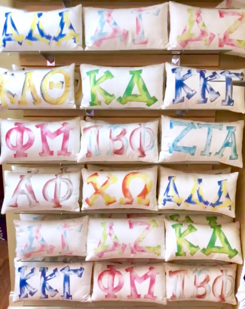 May include: A collection of 12 white pillow covers with colorful tie-dye designs featuring Greek letters. The letters include:  &Alpha;&Delta;&Pi;, &Kappa;&Delta;&Kappa;, &Kappa;&Kappa;&Gamma;, &Phi;&Mu;, &Tau;&Beta;&Phi;, &Zeta;&Tau;&Alpha;, &Alpha;&Phi;&Phi;, &Chi;&Omega;, &Delta;&Delta;&Delta;, &Delta;&Sigma;&Zeta;, &Kappa;&Delta;&Kappa;, &Kappa;&Kappa;&Gamma;, &Phi;&Mu;, &Tau;&Beta;&Phi;.