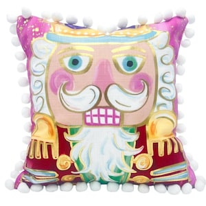 May include: A square decorative pillow featuring a colorful nutcracker illustration with a pink face, white mustache and beard, and a red uniform. The pillow is trimmed with white pom-poms along all four edges and has a vibrant, multi-colored design.