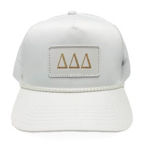 May include: White trucker hat with a mesh back and a curved brim. The hat features a rectangular patch with three gold-colored Greek letter deltas. A white rope detail accents the brim.