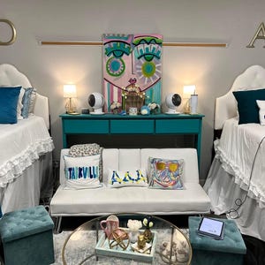 May include: Two twin beds with white bedding and blue and white pillows, a white futon with three pillows, a teal blue console table with two white lamps, and a glass coffee table with a tray of decorative items. The walls are painted a light gray and the room has a modern, eclectic style.
