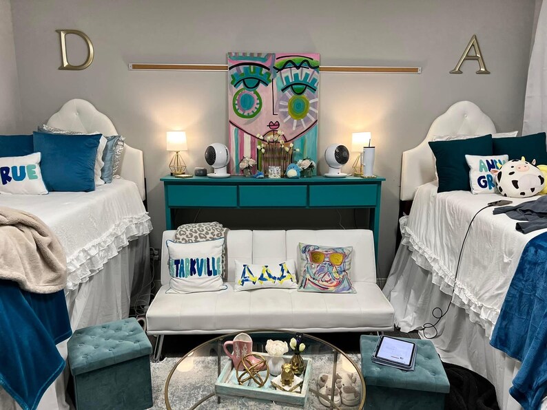 May include: Two twin beds with white bedding and blue and white pillows, a white futon with three pillows, a teal blue console table with two white lamps, and a glass coffee table with a tray of decorative items. The walls are painted a light gray and the room has a modern, eclectic style.