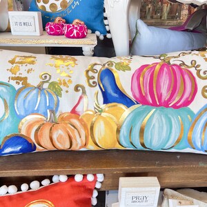 May include: A white cushion with a gold, blue, pink and orange pumpkin design. The pumpkins are painted with gold accents and have an autumnal theme.