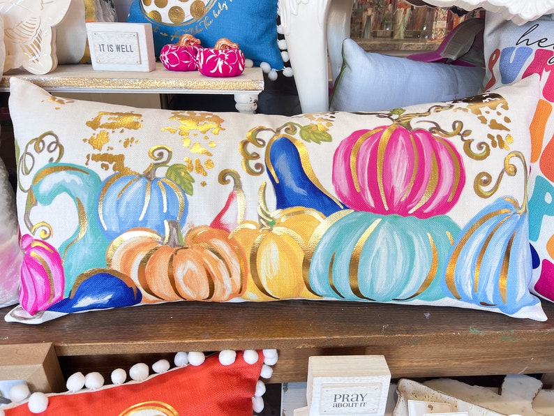 May include: A white cushion with a gold, blue, pink and orange pumpkin design. The pumpkins are painted with gold accents and have an autumnal theme.