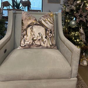 May include: A light gray armchair with a throw pillow featuring a gold and brown illustration of the nativity scene. The pillow has a gold star and a gold halo above the figures.