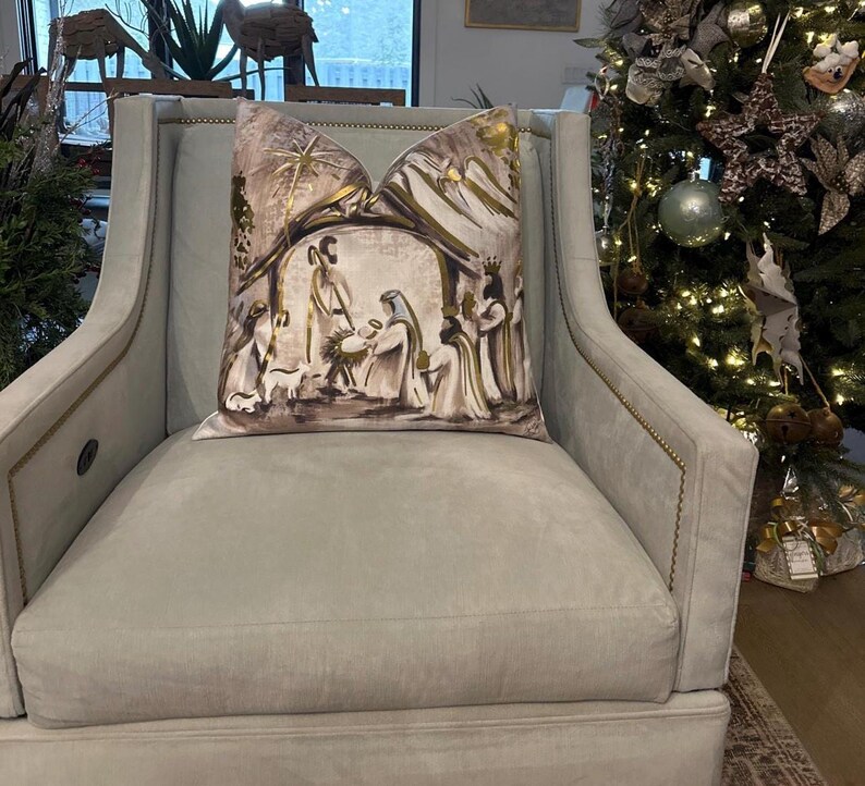 May include: A light gray armchair with a throw pillow featuring a gold and brown illustration of the nativity scene. The pillow has a gold star and a gold halo above the figures.