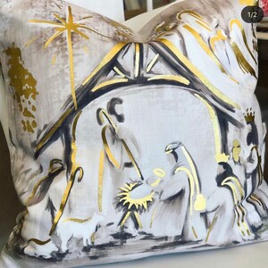 May include: A white throw pillow with a gold and black illustration of the nativity scene. The illustration features a star, a stable, and three figures.