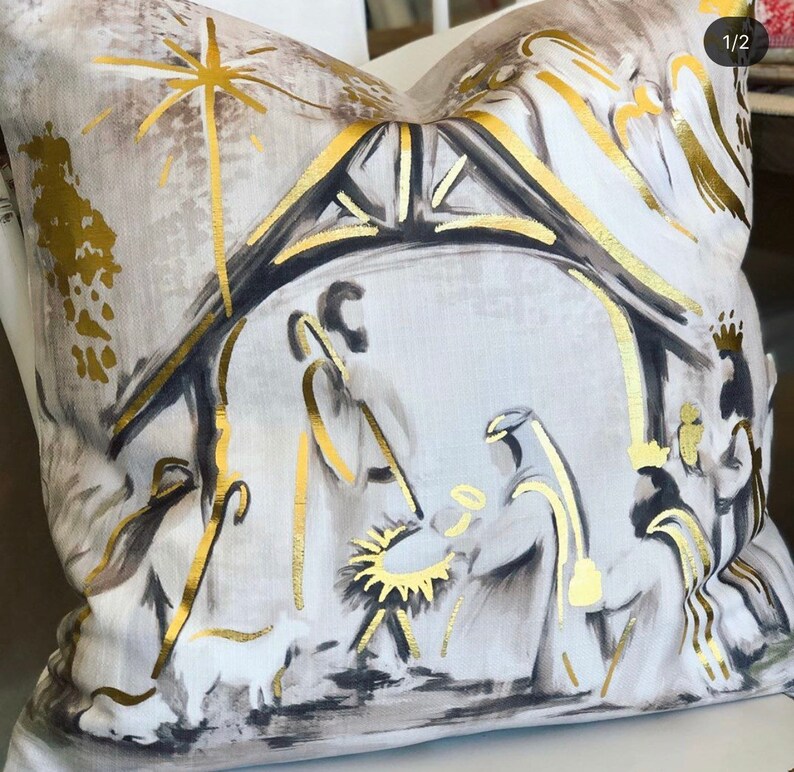 May include: A white throw pillow with a gold and black illustration of the nativity scene. The illustration features a star, a stable, and three figures.