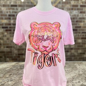 May include: Pink t-shirt with a gold glitter tiger wearing star-shaped sunglasses and the word "Tigers" in gold glitter.