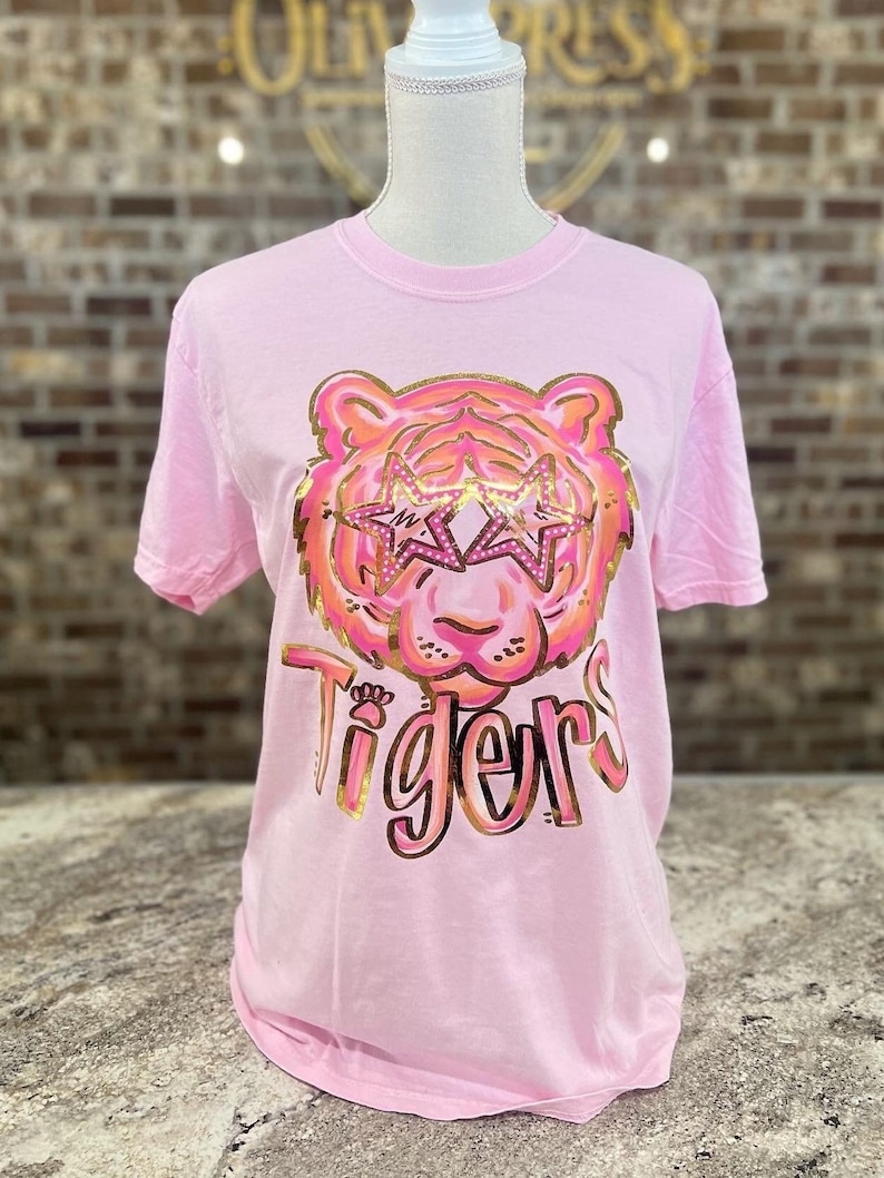 May include: Pink t-shirt with a gold glitter tiger wearing star-shaped sunglasses and the word "Tigers" in gold glitter.