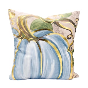 May include: A decorative throw pillow with a blue and gold pumpkin design. The pumpkin is painted in a light blue color with gold accents. The background is a light beige color.