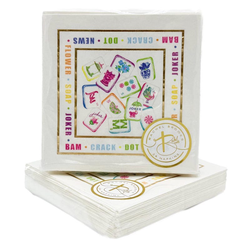 May include: White square napkins with a gold border and colorful illustrations. The napkins feature a gold seal with the text "RACHEL BROOKS 24 NAPKINS". The napkins have the words "FLOWER SOAP JOKER" and "BAM CRACK DOT NEWS" printed on them.