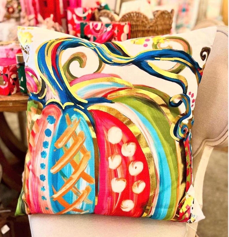 May include: A square throw pillow featuring a vibrant, multi-colored pumpkin illustration on a cream background. The pumpkin design incorporates bold strokes of blue, red, green, and gold, with decorative patterns including dots and cross-hatching.