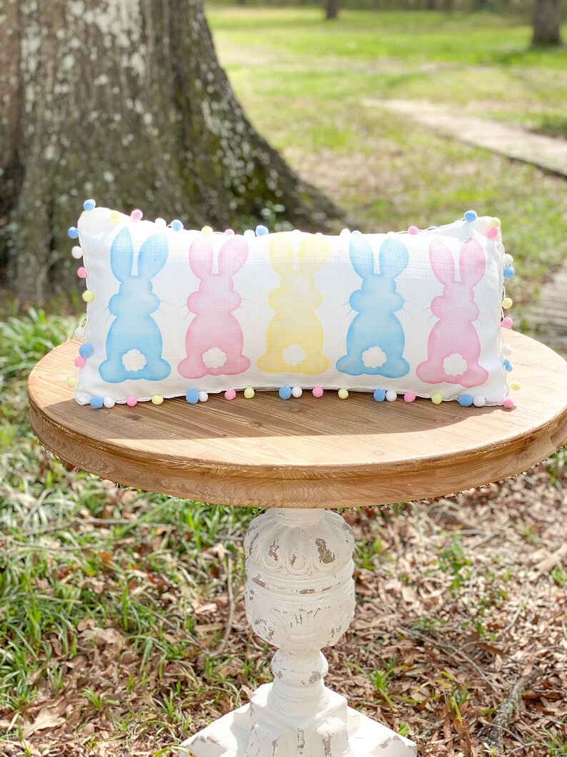 May include: A rectangular white pillow with pastel bunny silhouettes in blue, pink, and yellow. The pillow is trimmed with colorful pom-poms and rests on a round wooden table. The table is supported by a white, ornate pedestal.