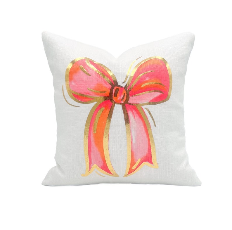 May include: White pillow with a pink and gold bow design. The bow is drawn in a whimsical style with a gold outline.