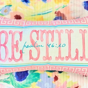 May include: A light green and pink pillow with a white border and a pink "BE STILL" inscription with the text "psalm 46:10" in blue underneath.