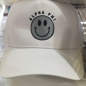 May include: White mesh trucker hat with a black embroidered smiley face and the text "ALPHA PHI" above it.