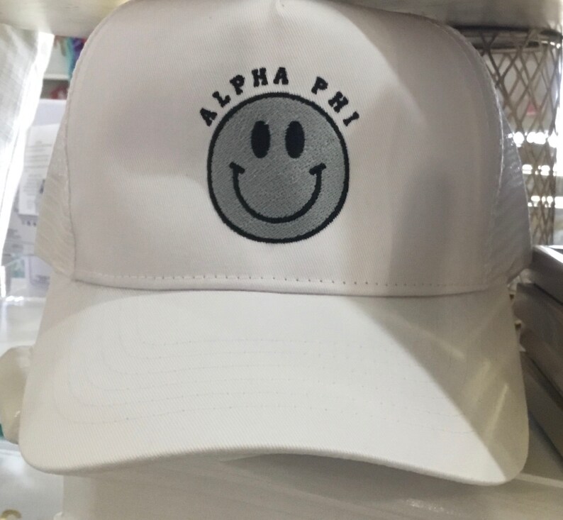 May include: White mesh trucker hat with a black embroidered smiley face and the text "ALPHA PHI" above it.