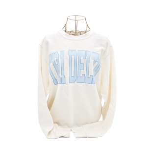 Op de afbeelding: Witte longsleeve waffle-trui met een blauwe "CHI DELTA" print.
