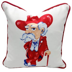 May include: White square pillow with a red border featuring a cartoon illustration of a man in a red suit and hat, leaning on a cane. The man has a white mustache and eyebrows, and a blue bow tie.