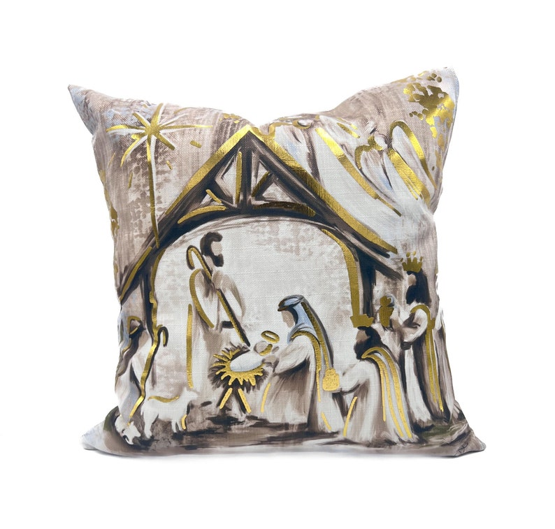 May include: A beige throw pillow with a gold and brown printed design of the nativity scene. The pillow features a star, a manger, and figures of Mary, Joseph, and the baby Jesus.