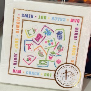 May include: A stack of white paper napkins with a gold border and colorful illustrations. The napkins feature various designs, including flowers, a peacock, and the word "Joker." The brand name "Rachel Brooks" is visible on a gold sticker.