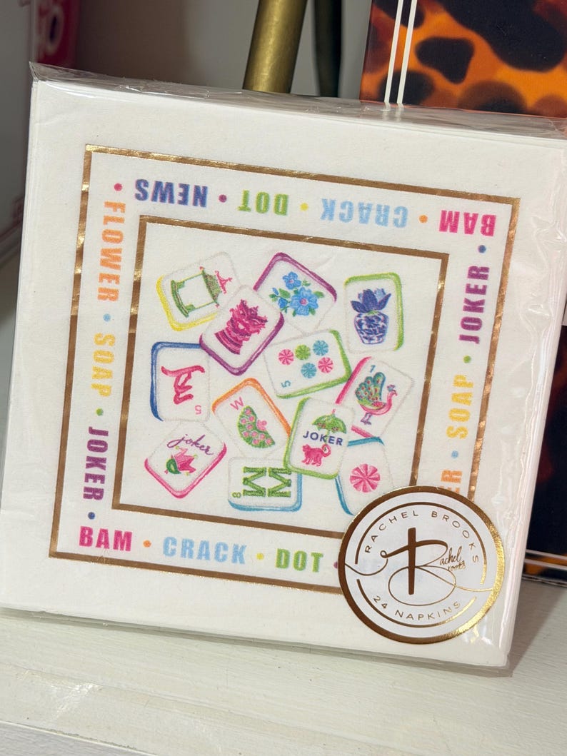 May include: A stack of white paper napkins with a gold border and colorful illustrations. The napkins feature various designs, including flowers, a peacock, and the word "Joker." The brand name "Rachel Brooks" is visible on a gold sticker.
