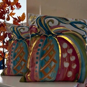 May include: Two square throw pillows featuring a painted pumpkin design on a white background. The pumpkins are decorated with vibrant teal, pink, and orange brushstrokes, accented by metallic gold outlines and details throughout the surface.