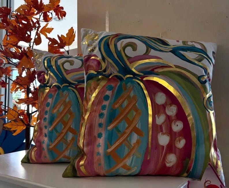 May include: Two square throw pillows featuring a painted pumpkin design on a white background. The pumpkins are decorated with vibrant teal, pink, and orange brushstrokes, accented by metallic gold outlines and details throughout the surface.
