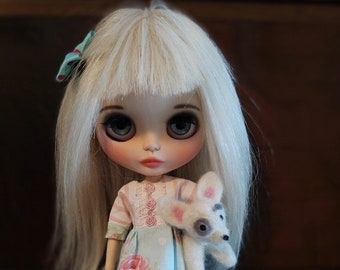 Puede incluir: Una muñeca con cabello largo y liso de color blanco y un lazo azul cerceta, que viste un vestido azul claro con estampado de rosas rosas, lunares blancos y corpiño a rayas. La muñeca sostiene un pequeño animal de fieltro blanco con manchas grises.