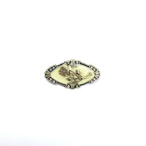 以下が含まれることがあります： クリーム色の中心と花の模様が特徴的な、楕円形のヴィンテージブローチ。ブローチは、縁に小さな虹色のクリスタルが付いた銀色の金属フレームで囲まれています。花の模様は浮き彫りになっており、立体感を与えています。