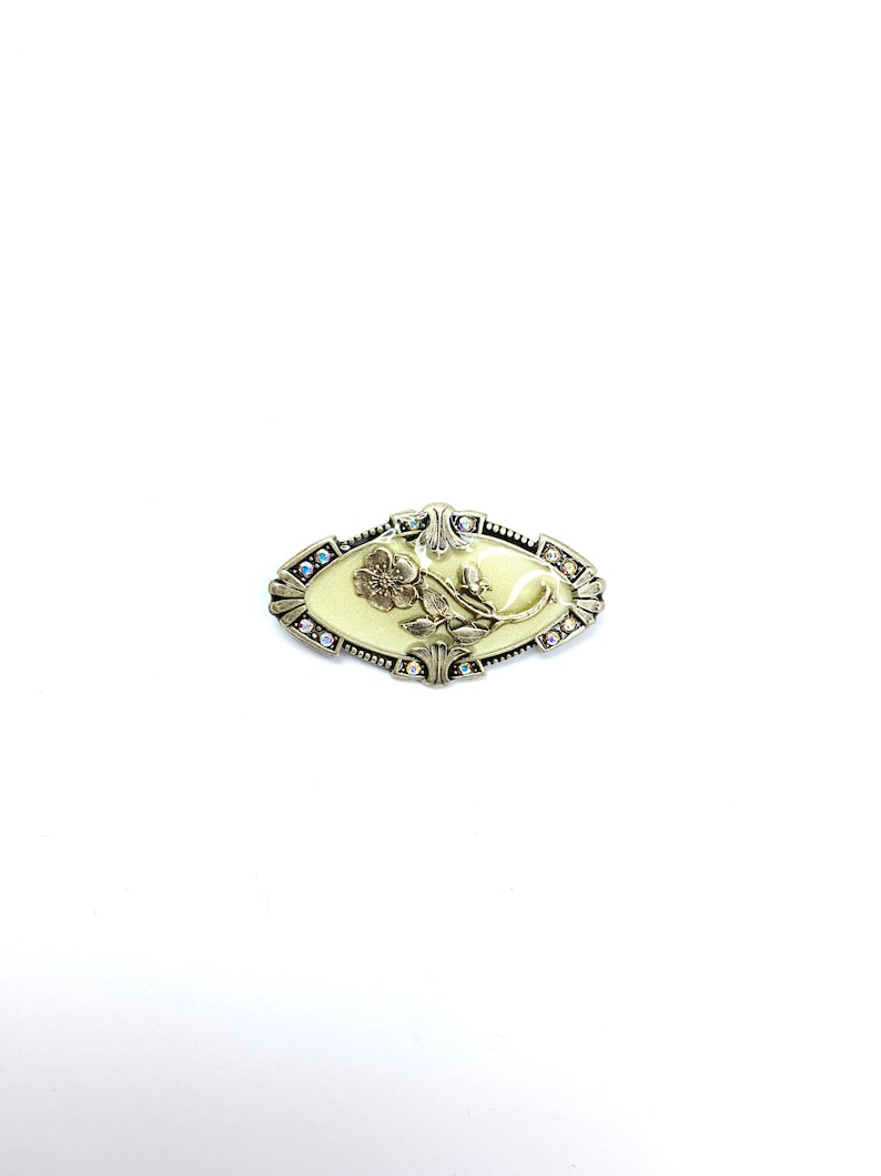 以下が含まれることがあります： クリーム色の中心と花の模様が特徴的な、楕円形のヴィンテージブローチ。ブローチは、縁に小さな虹色のクリスタルが付いた銀色の金属フレームで囲まれています。花の模様は浮き彫りになっており、立体感を与えています。