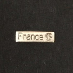 以下が含まれることがあります： クラシックなフォントで「France」の文字が刻まれた、小さく長方形のシルバー調の金属タグ。タグには小さなシンボルも付いています。タグは暗い、おそらく黒の背景に配置されています。